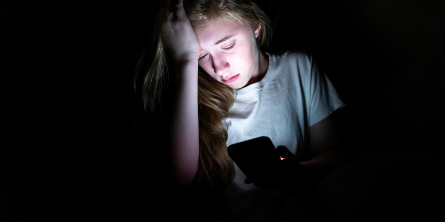 Ein Teenager sitzt im Dunklen und schaut etwas auf dem hell erläuchteten Handy an.