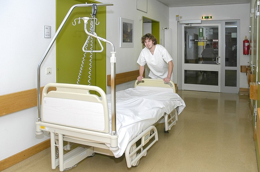 Ein Krankenhelfer schiebt ein Krankenbett einen Gang im Krankenhaus entlang.